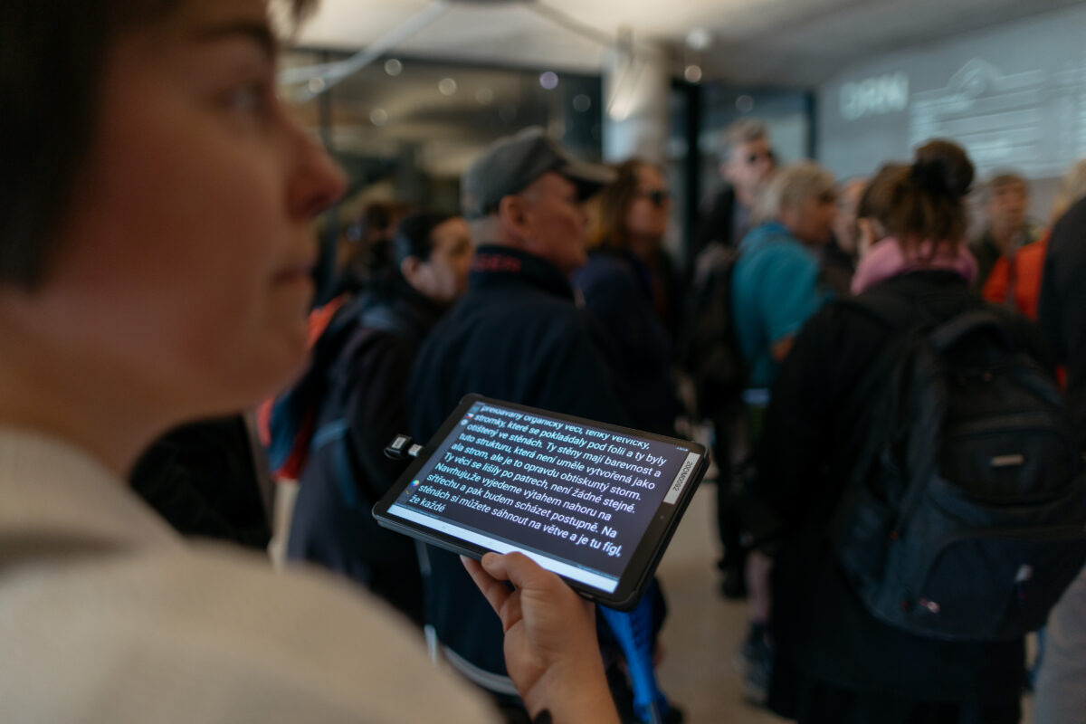 V popředí rozostřená postava držící tablet, na kterém běží přepis komentované prohlídky v&nbsp;prostorách CAMP.
